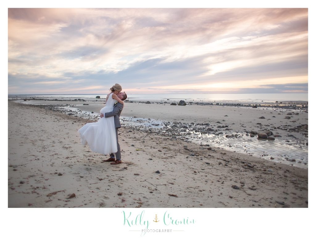A man lifts his bride up for a kiss on the shore near The Dennis Inn, captured by Kelly Cronin Photography