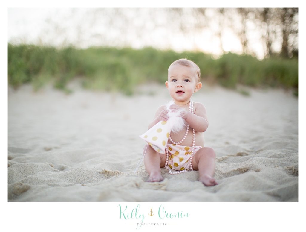 A baby girl sits on the beach and plays with a birthday hat for her cake smash session. 