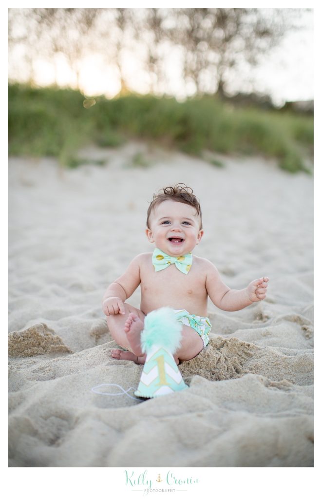 A baby boy sits on the beach and plays with a birthday hat. 