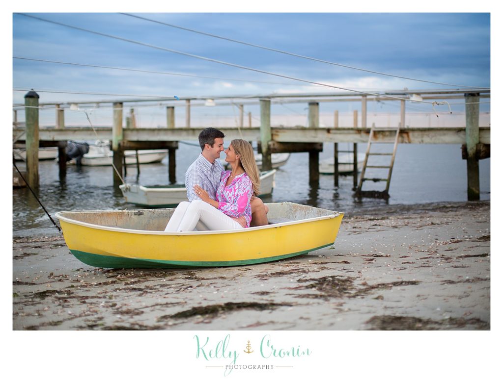 An adult couple sit in a little yellow boat on the shore. 