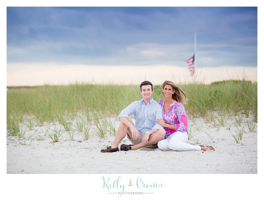 A couple sit in the sand in front of a grassy patch on the beach. 