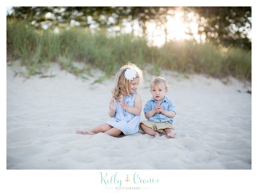 Two young children sit in the sand and clap their hands. 