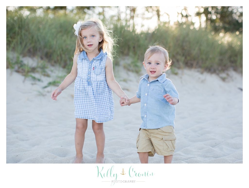 Two children hold hands as they walk along the beach. 
