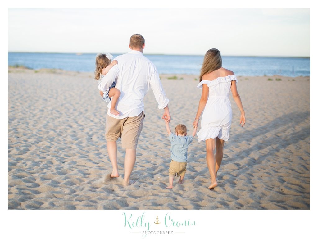 A family of four walk along the beach while holding hands.