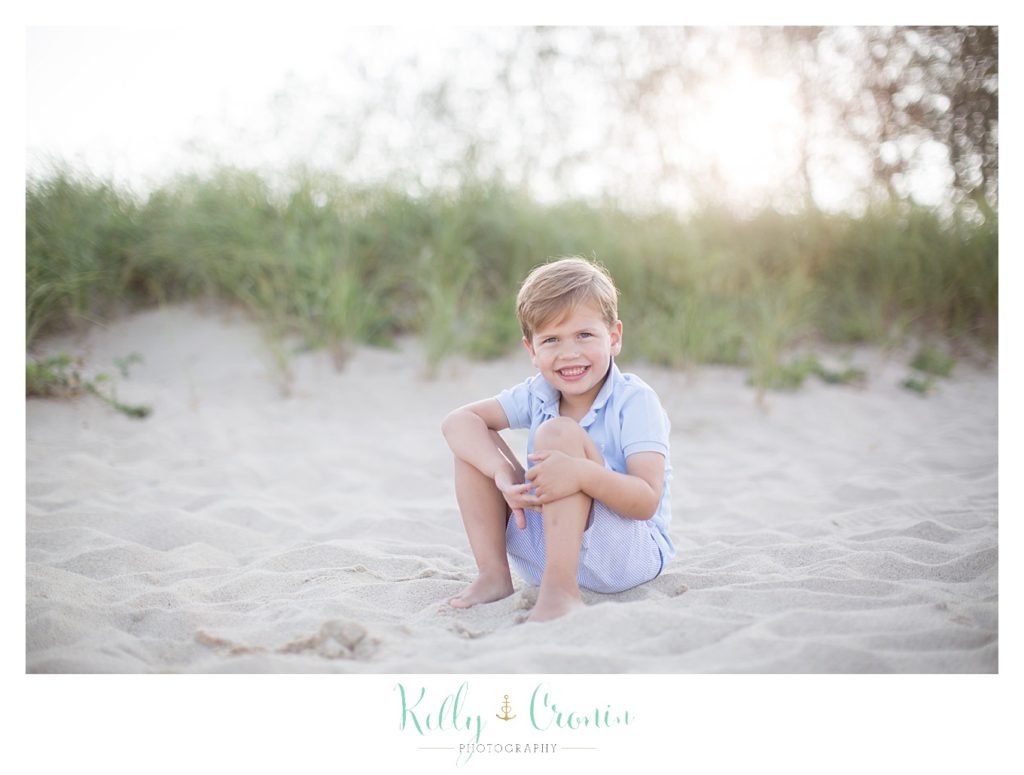 A boy sits in the sand holding his knees smiling. 
