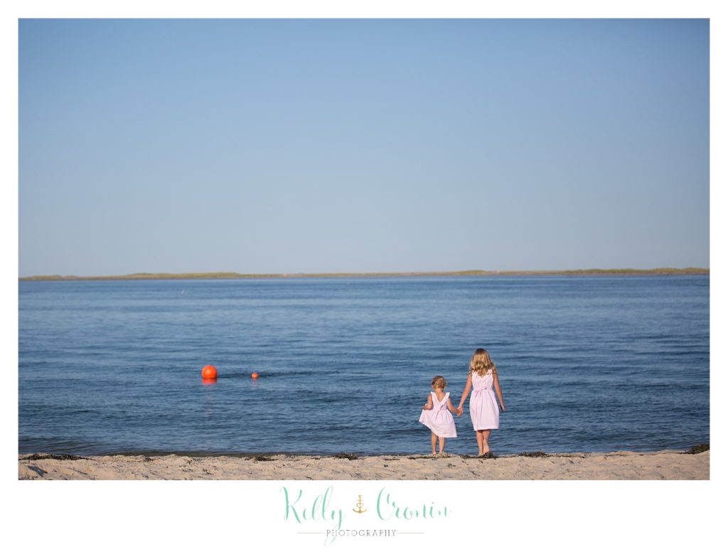 Two sisters stand at the shoreline looking at the water. 