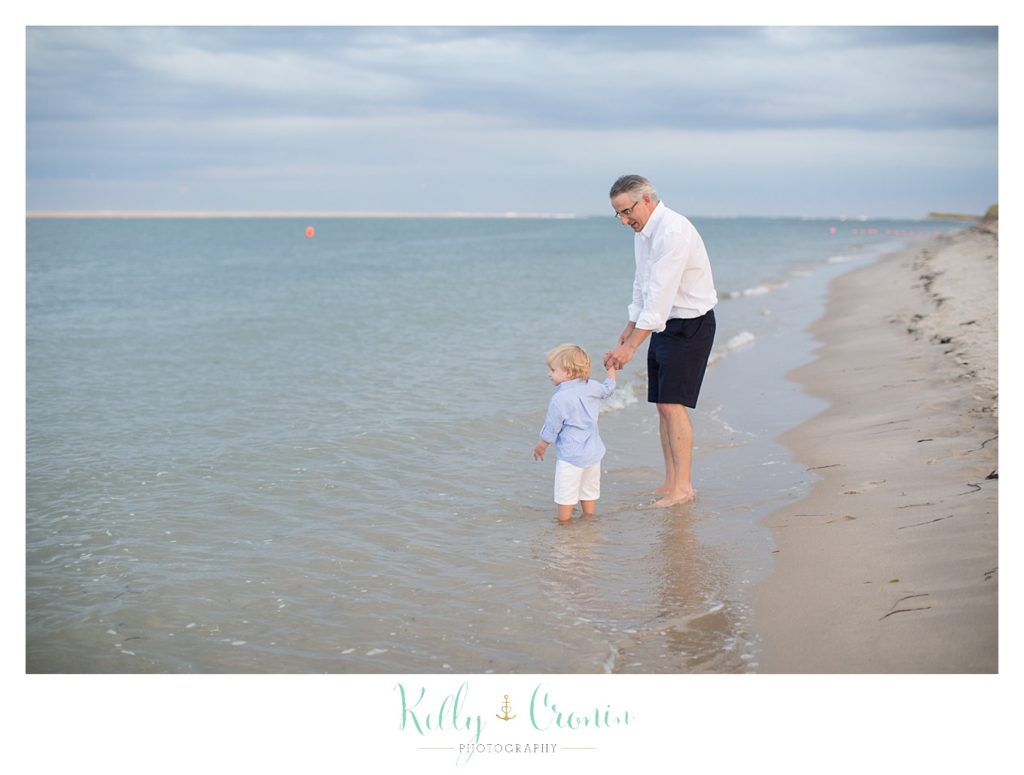 A man holds his son's hand as he walks through the waves. 