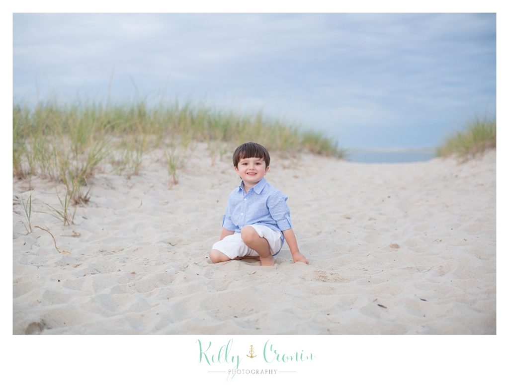 A boy is sitting on the beach and digging his hands into the sand. 