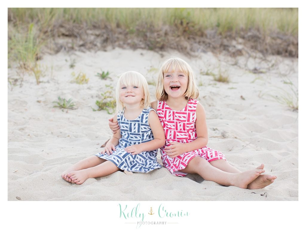 Two blonde little girls sit in the sand and laugh. 