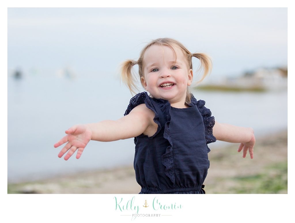 A toddler with pigtails twirls around on the beach. 