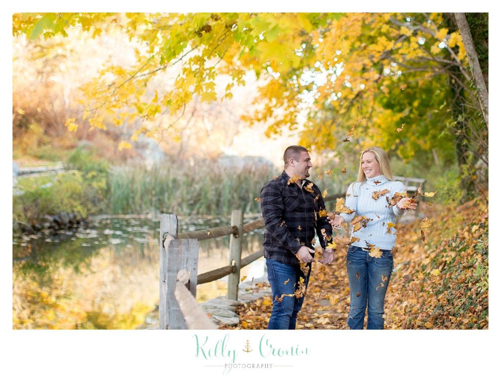 An engaged couple hold hands and walk among fallen leaves. 