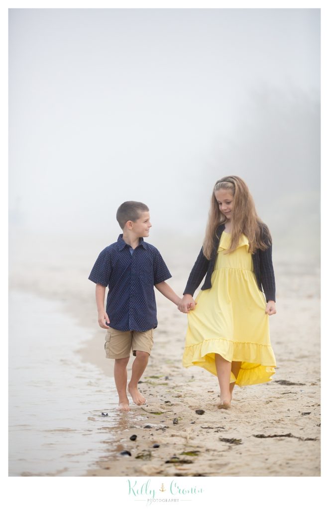 Siblings hold hands and walk on the sandy beach together. 