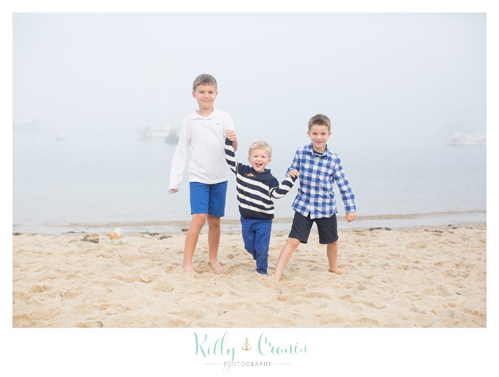 Three small boys stand in a line, holding hands on the beach. 
