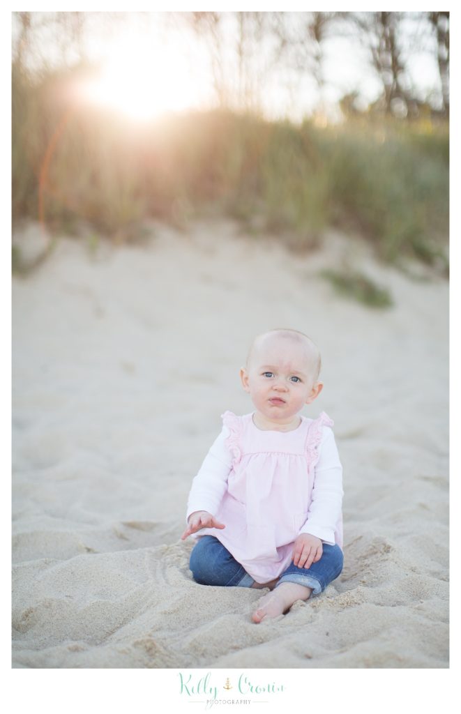 A blonde baby girl sits on the beach. 