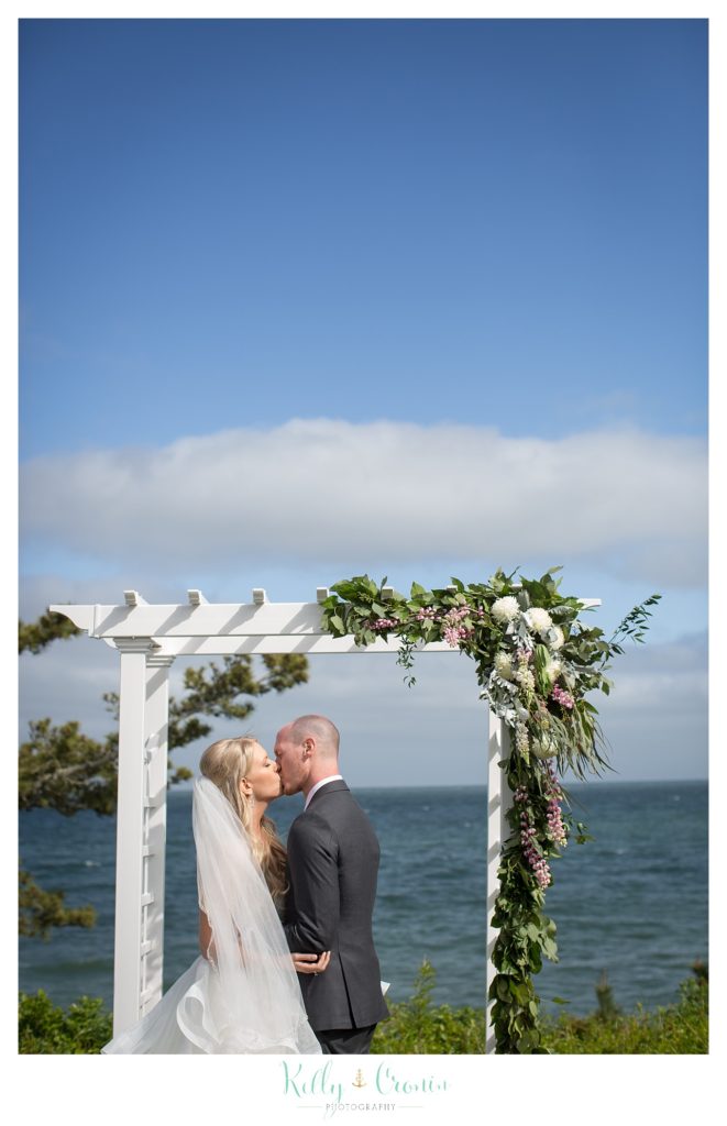 A bride and groom stand under an arch with flowers. 