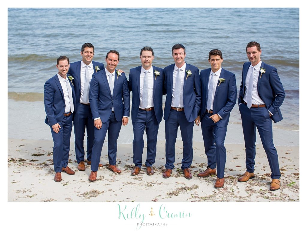 A groom's party stands in their tuxedos on the beach. 