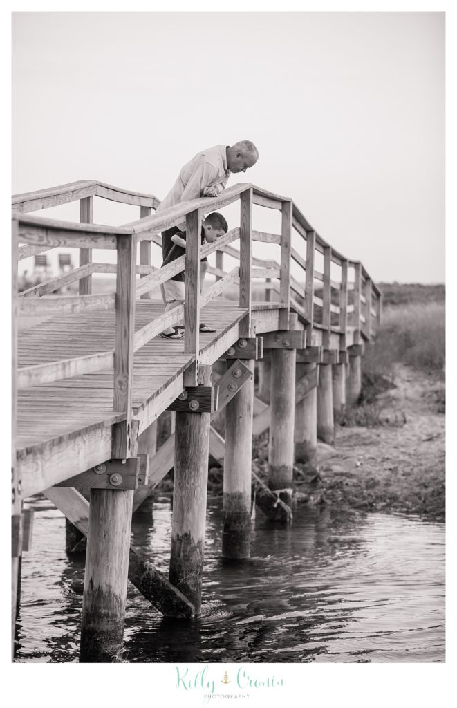 A boy and his grandpa lean over a bridge to look at the water. 