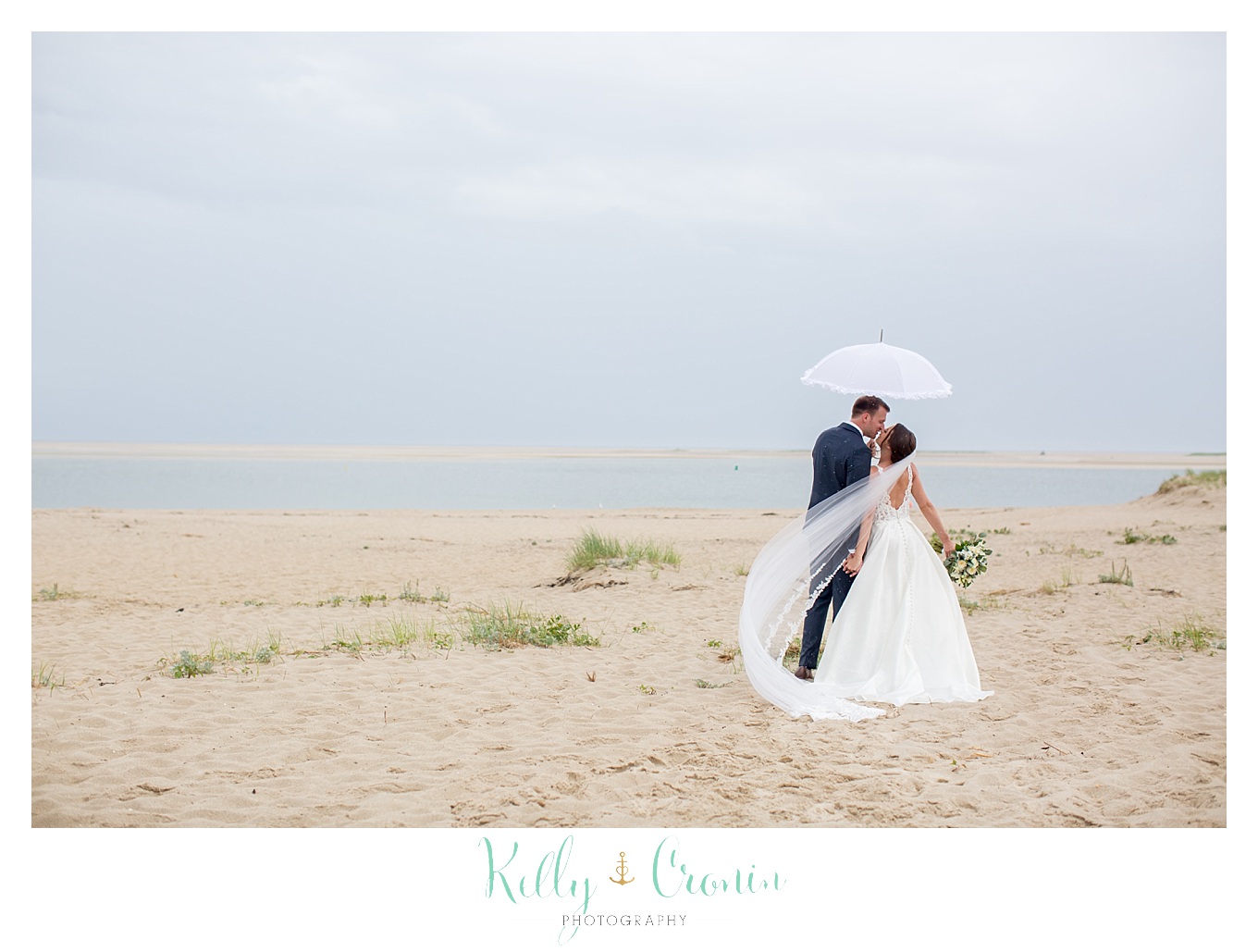 A bride and groom walk along the beach under an umbrella.