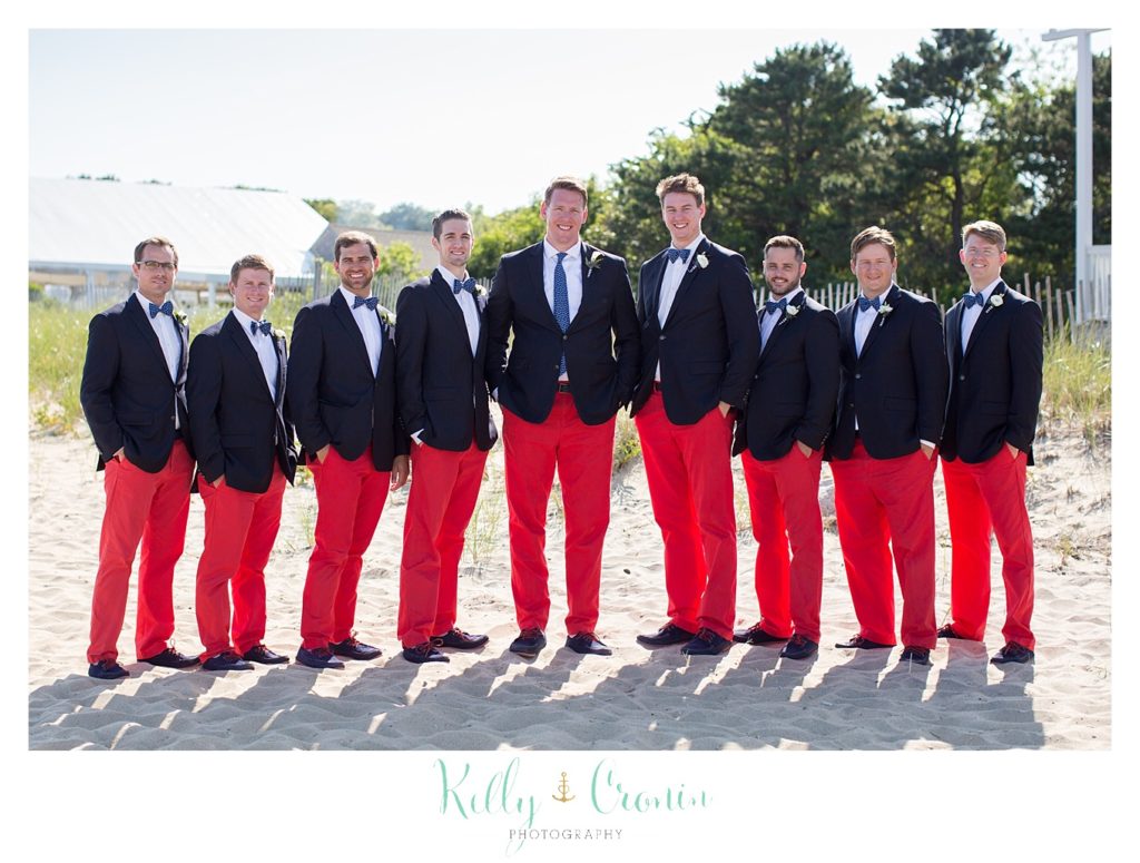 Groomsmen stand in the sand on the beach.