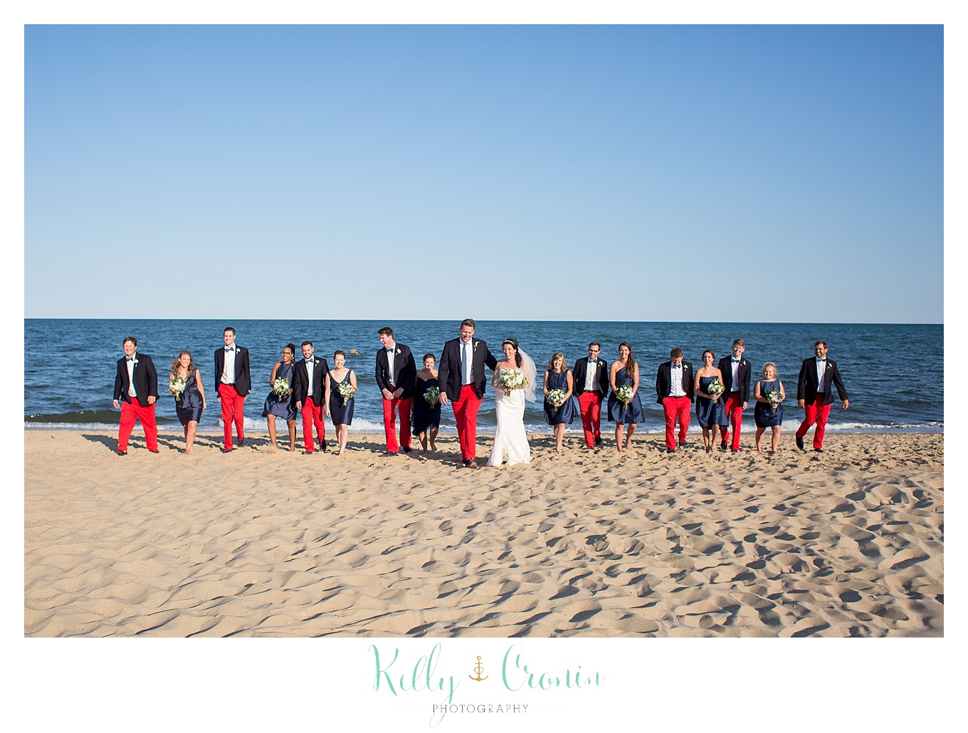 A wedding party stands in a line along the shoreline.