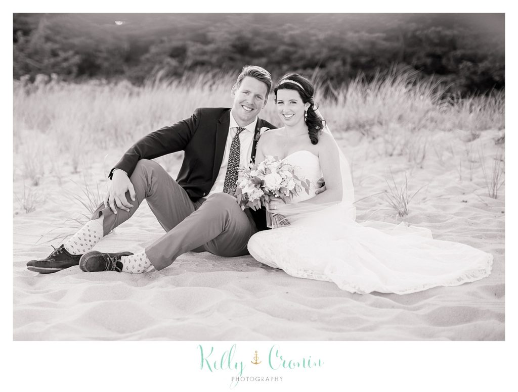 A bride and groom sit in the sand on the beach.