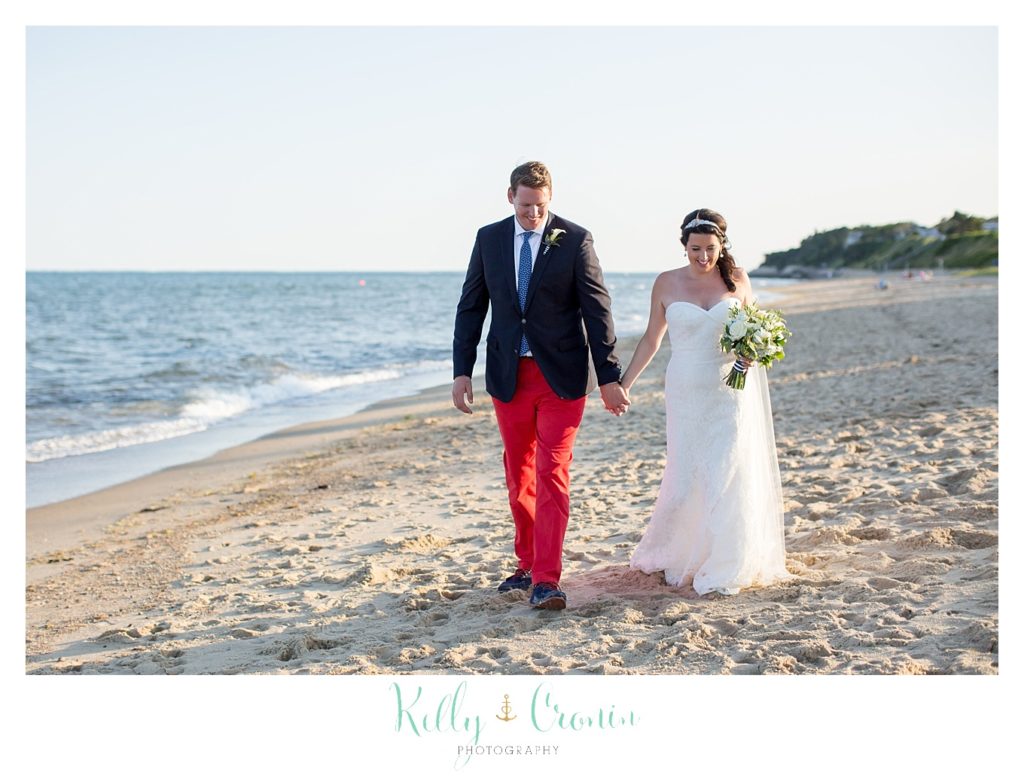 A bride and groom hold hands and walk down the beach.