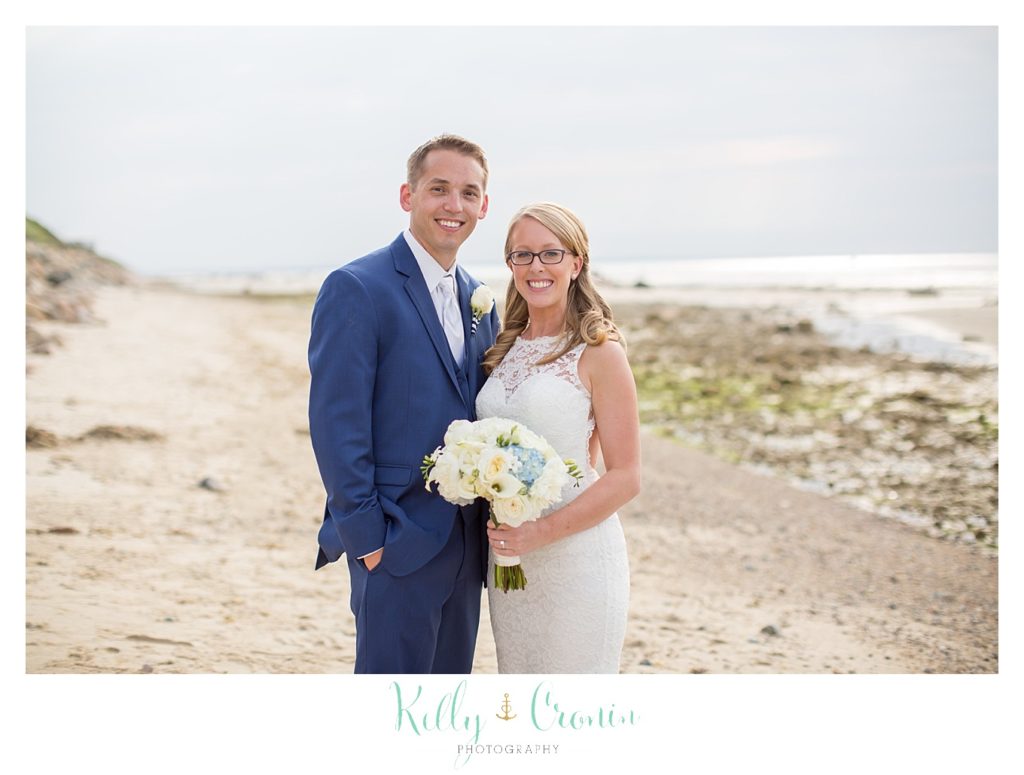 A bride and groom stand on a sandy beach together. 