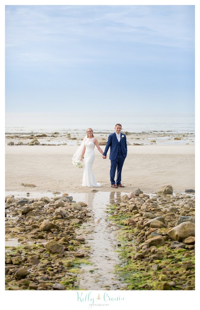 A bride and groom walk on the beach together. 