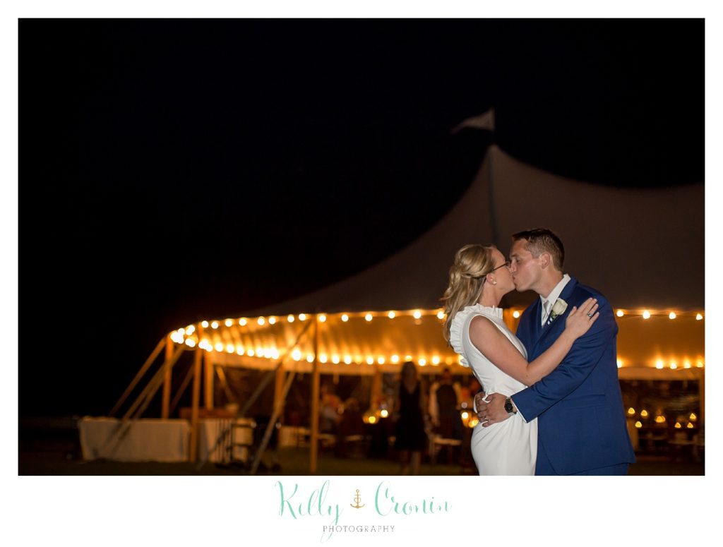 A bride and groom kiss in front of a lighted wedding tent. 