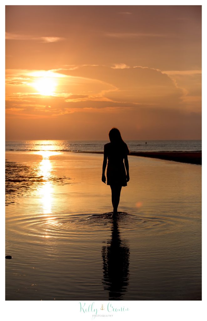 A woman walks along the shore as the sun sets. 