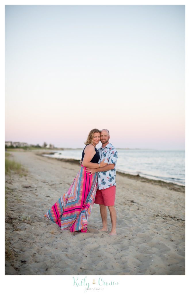 A woman hugs her husband while they stand in the sand on the beach. 