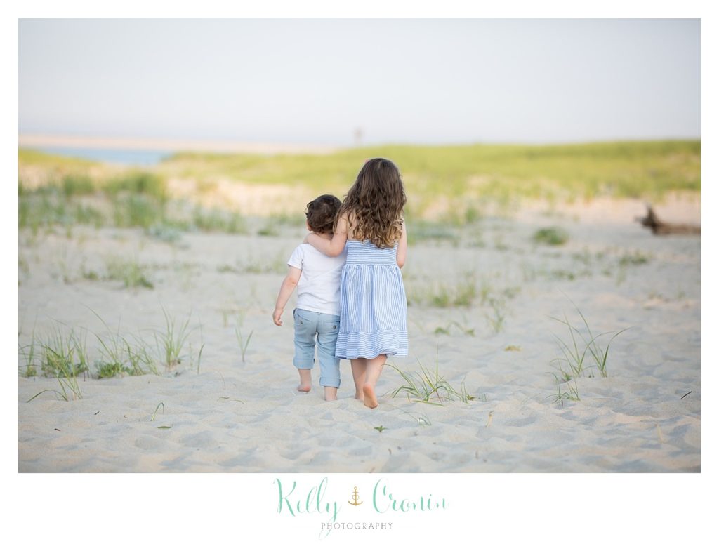 A little girl puts her arm around her baby brother as they walk along the beach. 