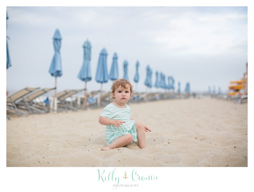 A toddler sits on the beach in front of umbrellas. 