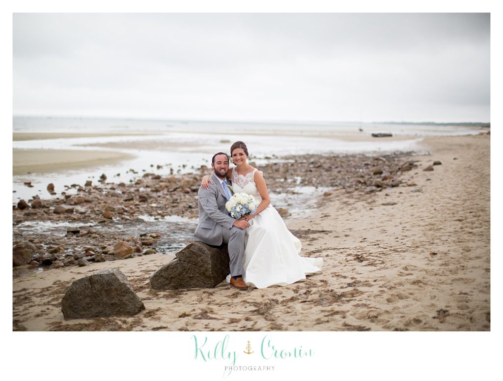 A bride and groom sit on a rock in front of the ocean. 