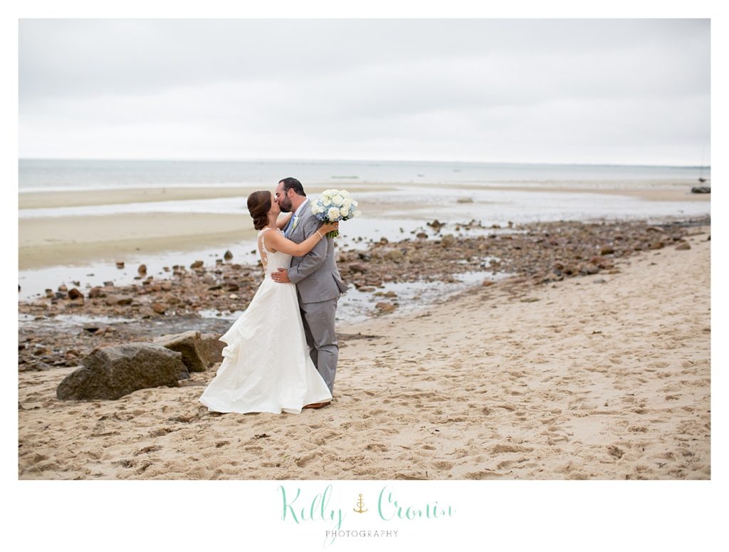 A groom kisses his bride in front of the shoreline. 