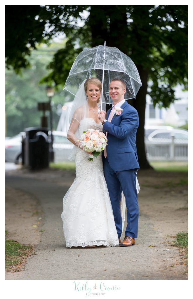 A bride and groom stand under an umbrella as the rain comes down. 
