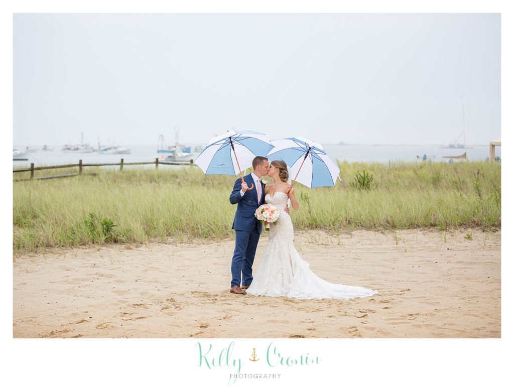 A bride and groom kiss under two beach umbrellas. 