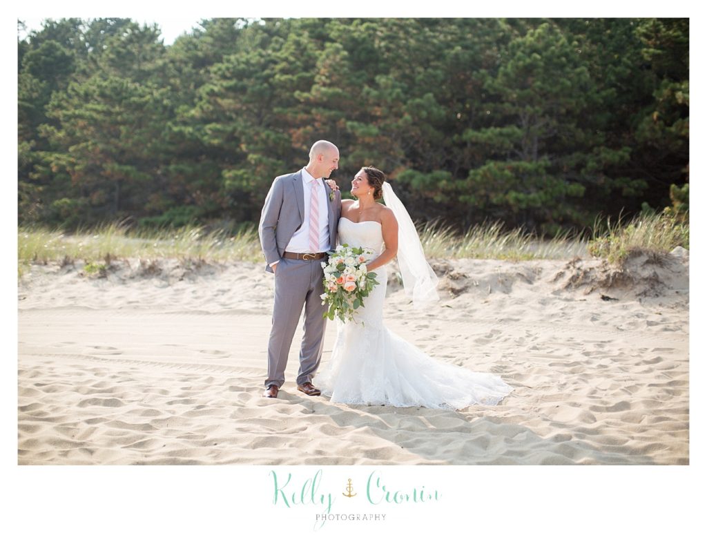A bride and groom stand in the sand on the beach. 