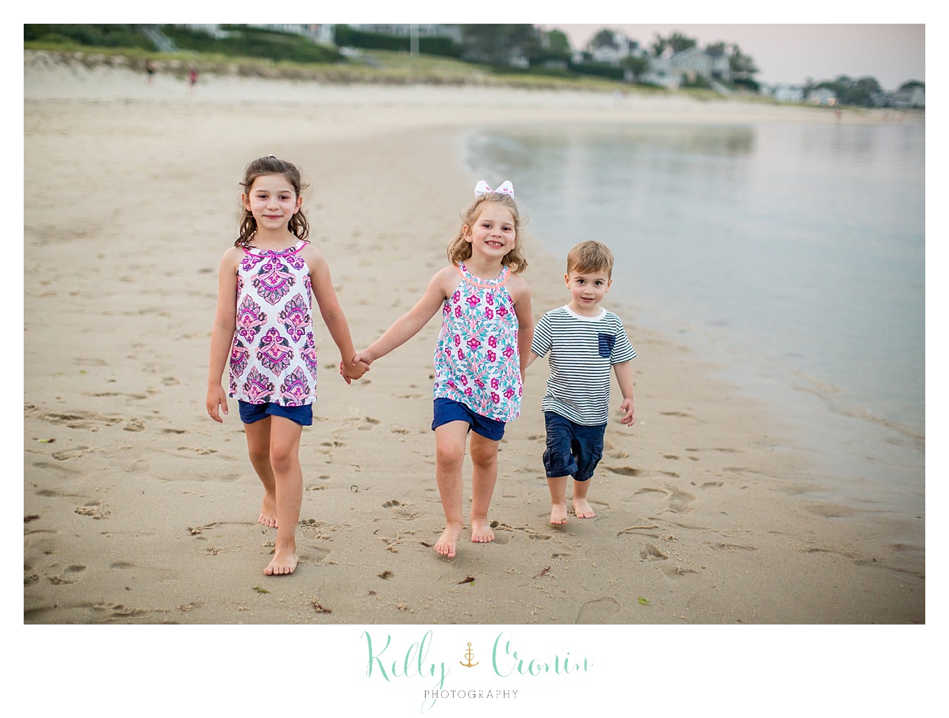 Three siblings hold hands while walking in the sand.