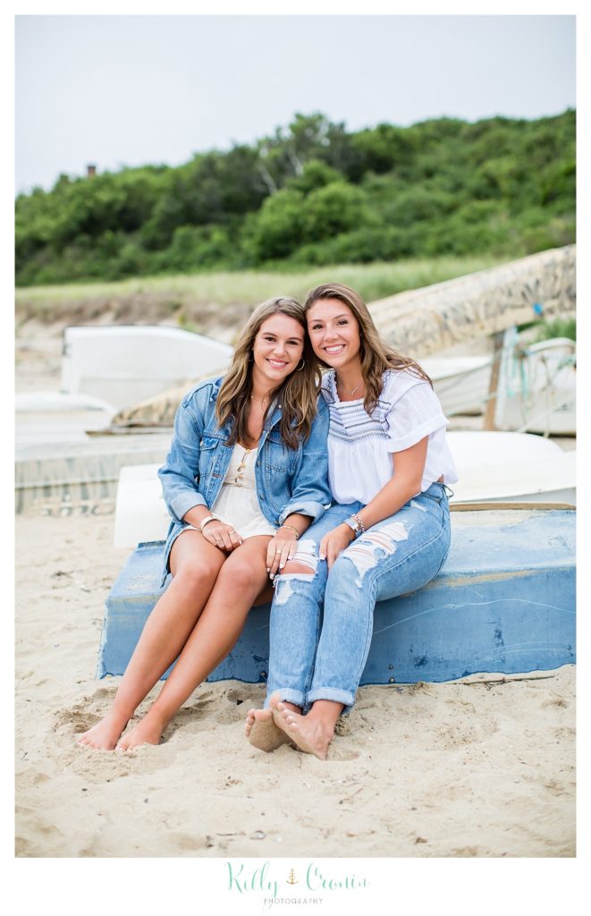 Two sisters sit on a stranded boat on the beach. 