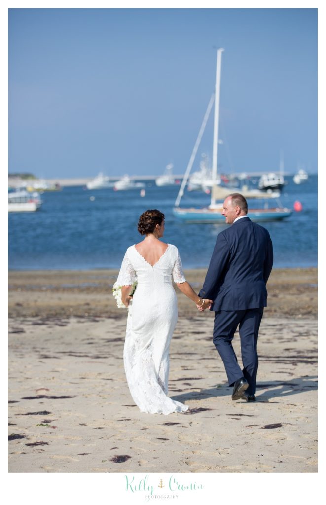 A bride and groom walk down a beach together. 