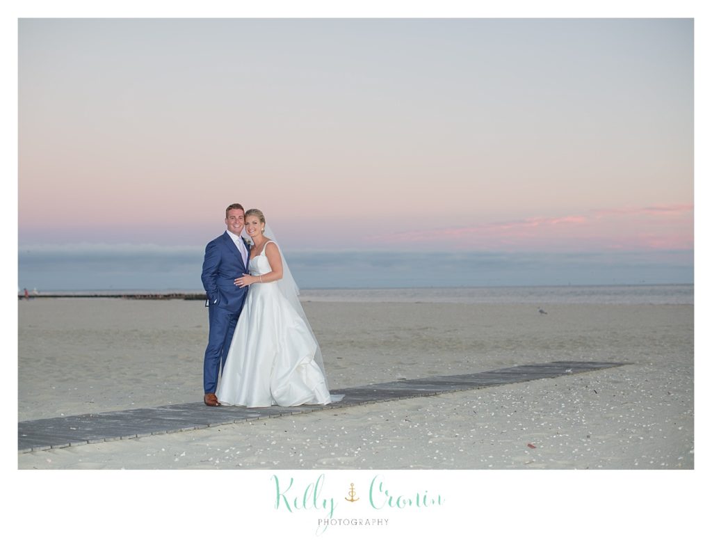 A bride and groom hug on the beach in front of a sunset. 