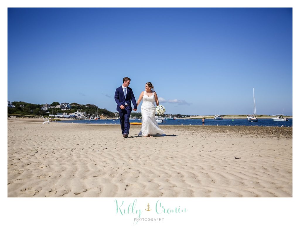 A bride and groom walk in the sand on the beach. 