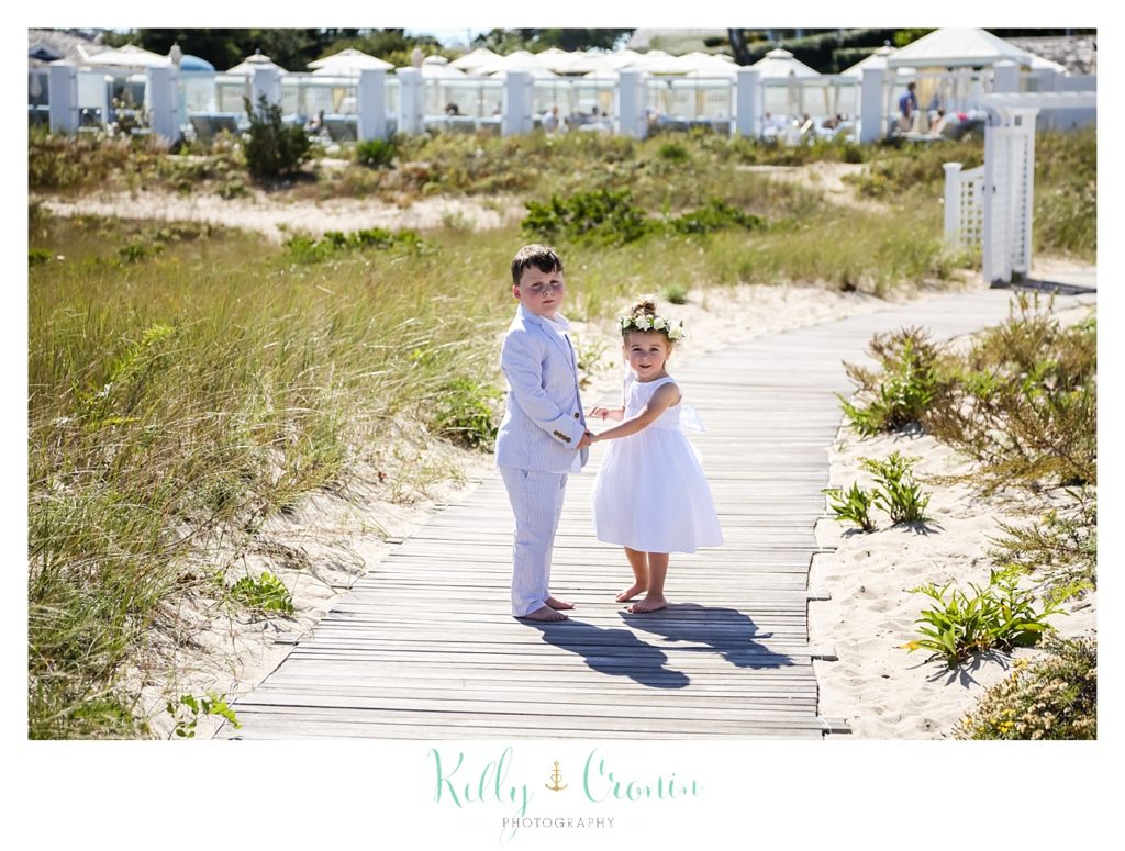 A flower girl and ring bearer hold hands while walking on the beach. 