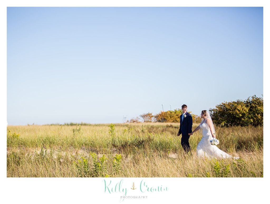 A bride and groom walk through tall grass on the beach. 