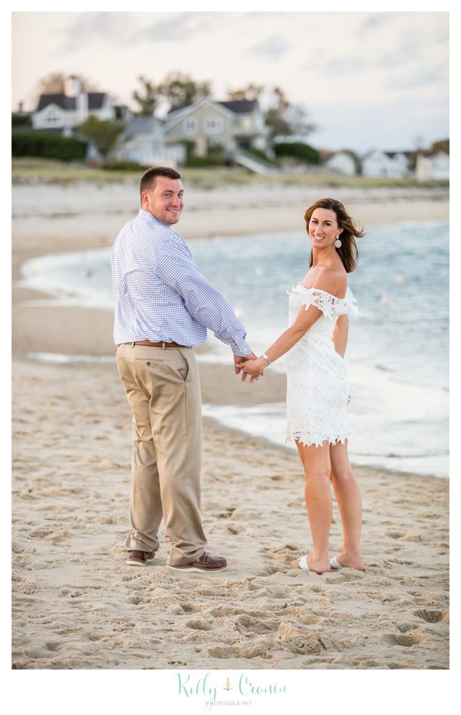 An engaged couple hold hands as they walk on the beach together. 