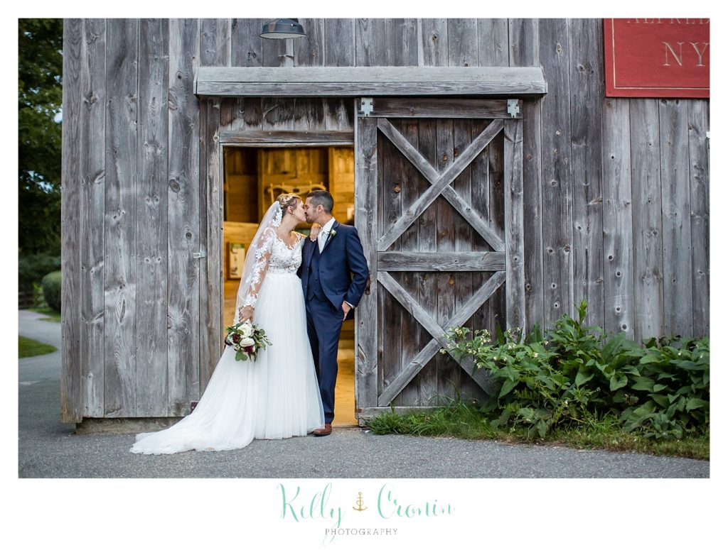 A bride and groom stand in front of a barn together. 