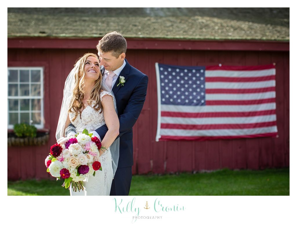 A bride and groom stand in front of an American flag. 
