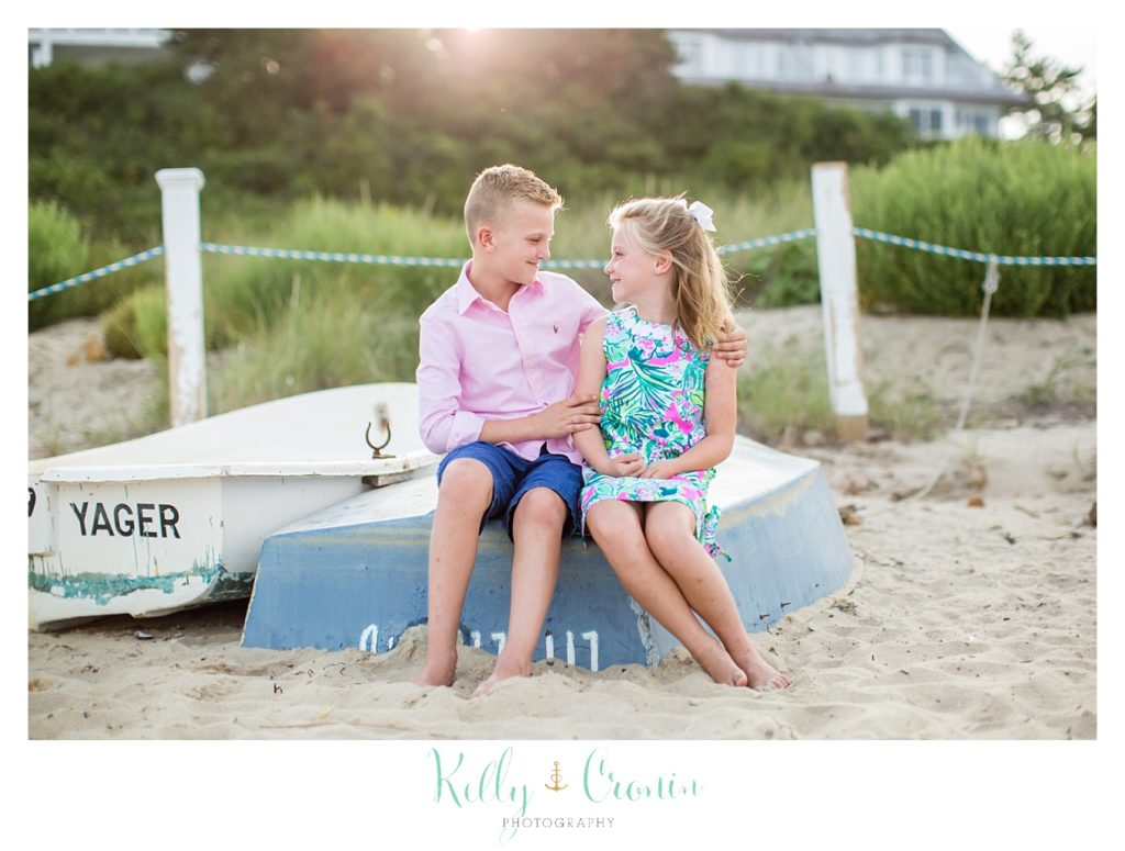 Two siblings sit on a boat on the sandy beach. 